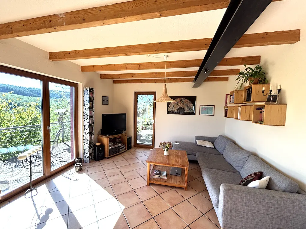 Living room with wood beams, tile floor, and gray sectional sofa. Balcony with green view visible through glass doors.