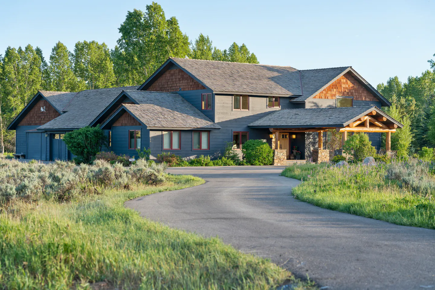 Exterior of a large, gray house with a wood porch and a winding driveway. Trees in the background.