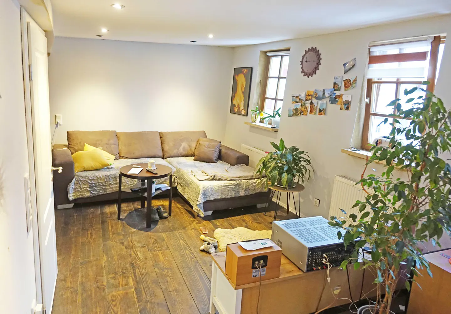 Living room with brown sectional sofa, round table, wood floors, and windows with natural light. Plants and decor add warmth.