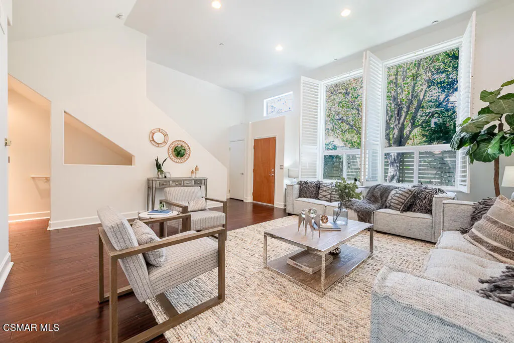 Bright living room with hardwood floors, white walls, and large windows. Two sofas face each other over a light rug and metal coffee table.