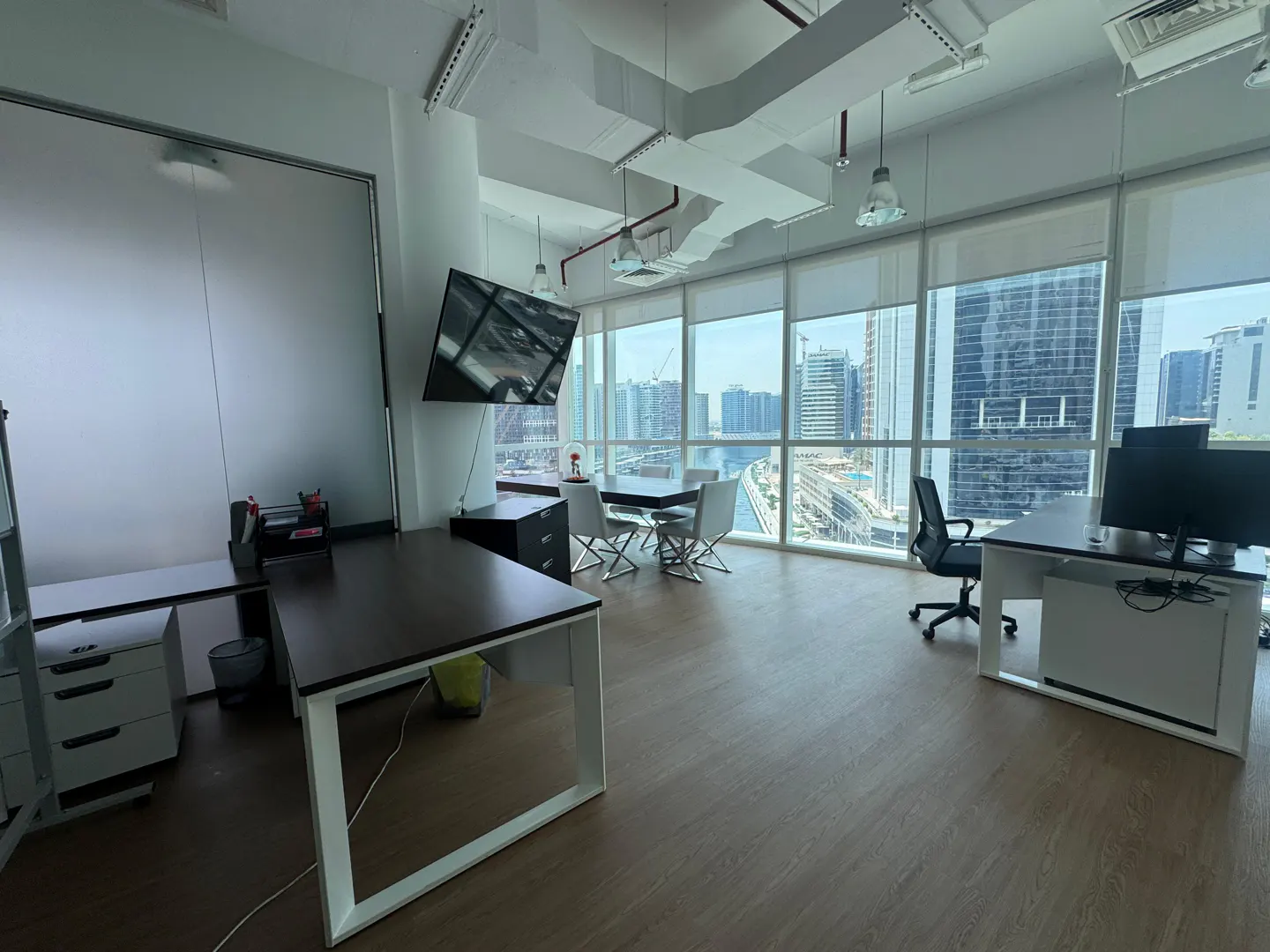 Bright office space with desks, chairs, and a large window view of a city skyline. A flat-screen TV is mounted on the wall.