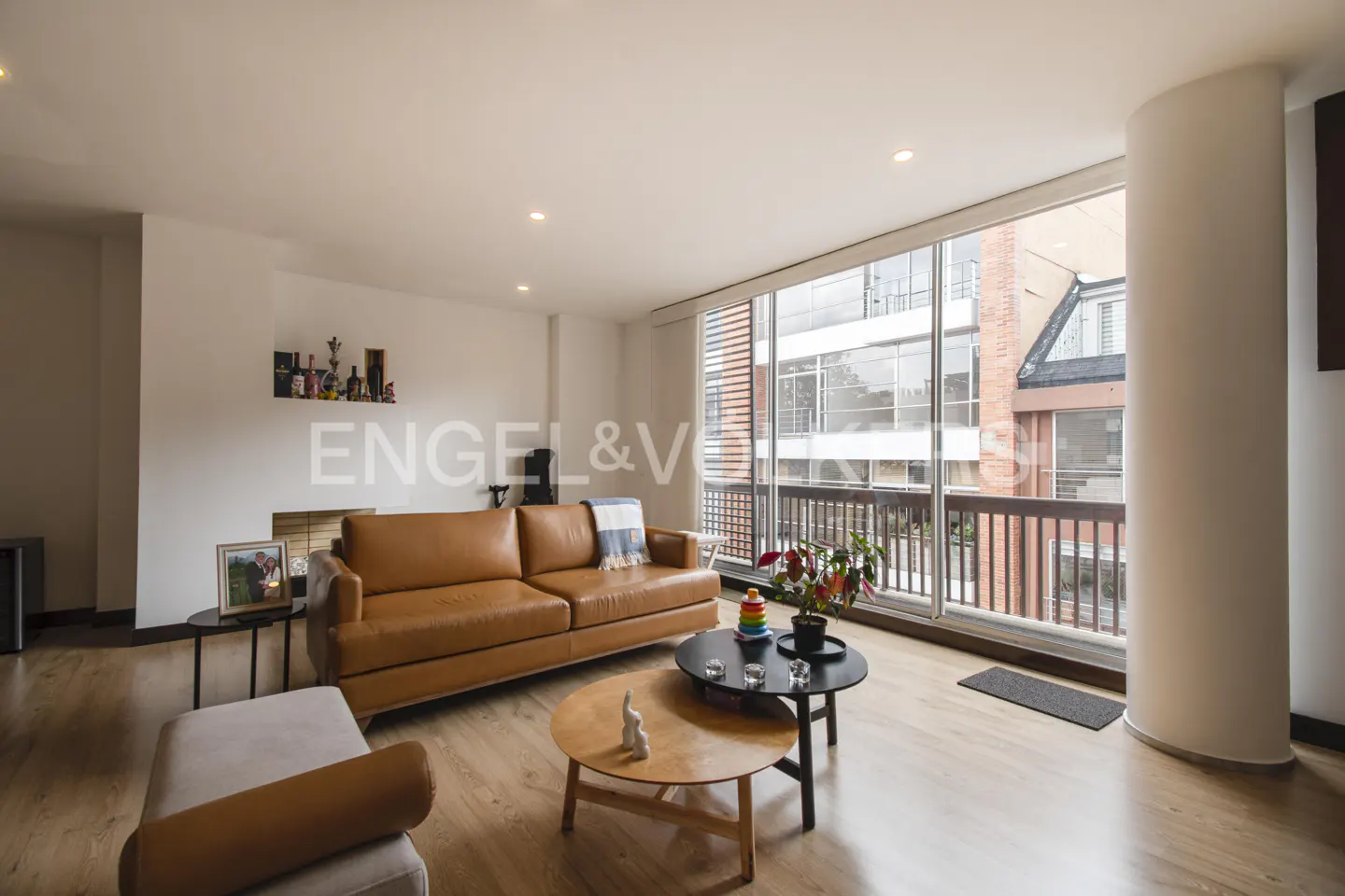 Bright living room with wood floors, a brown leather sofa, and a large window overlooking a balcony. Two round tables sit in front of the sofa.