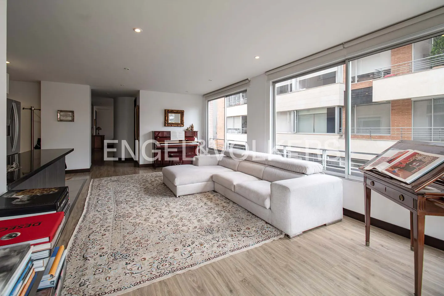 Bright living room with a white sectional sofa, patterned rug, and large windows overlooking a city building.
