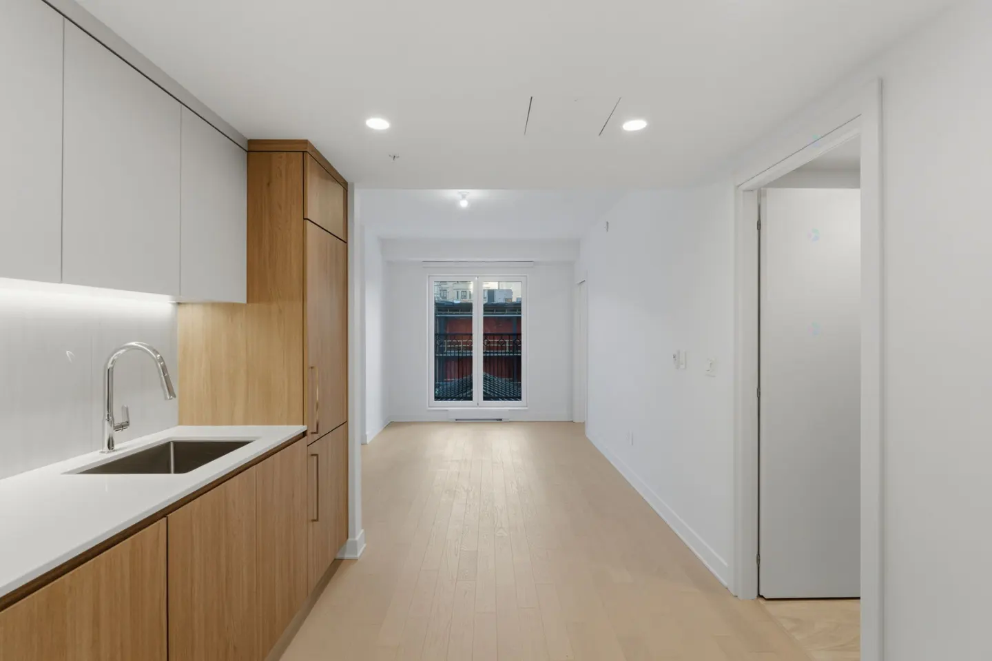 A bright, modern apartment interior with white walls, light wood floors, and a kitchen area with wood cabinets. A window shows a building exterior.