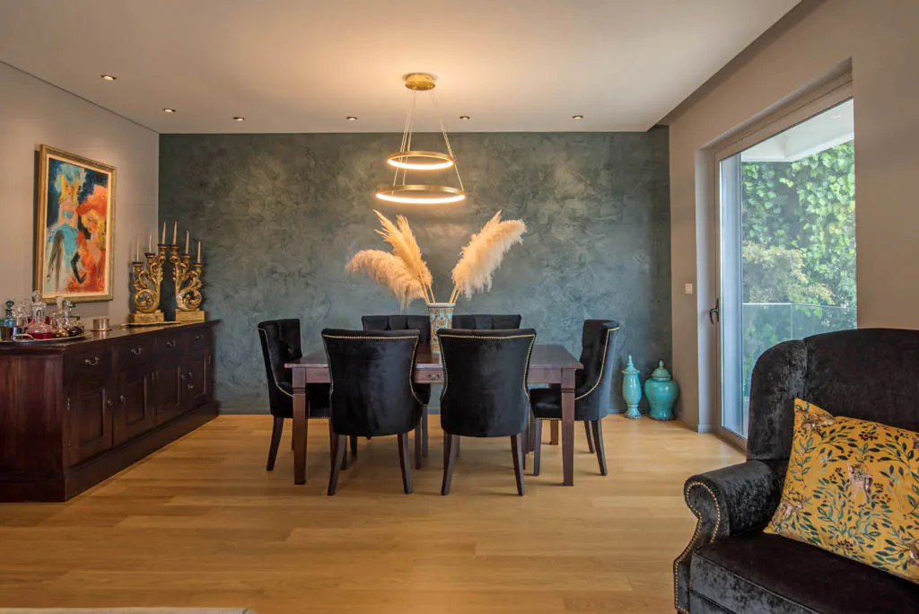 A dining room with a dark wood table, black velvet chairs, and a modern chandelier. A dark wood sideboard sits against a textured green wall.