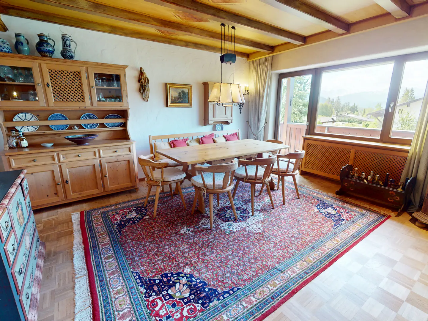 Rustic dining room with wooden beams, a large red patterned rug, wooden table, chairs, and a hutch with blue pottery.