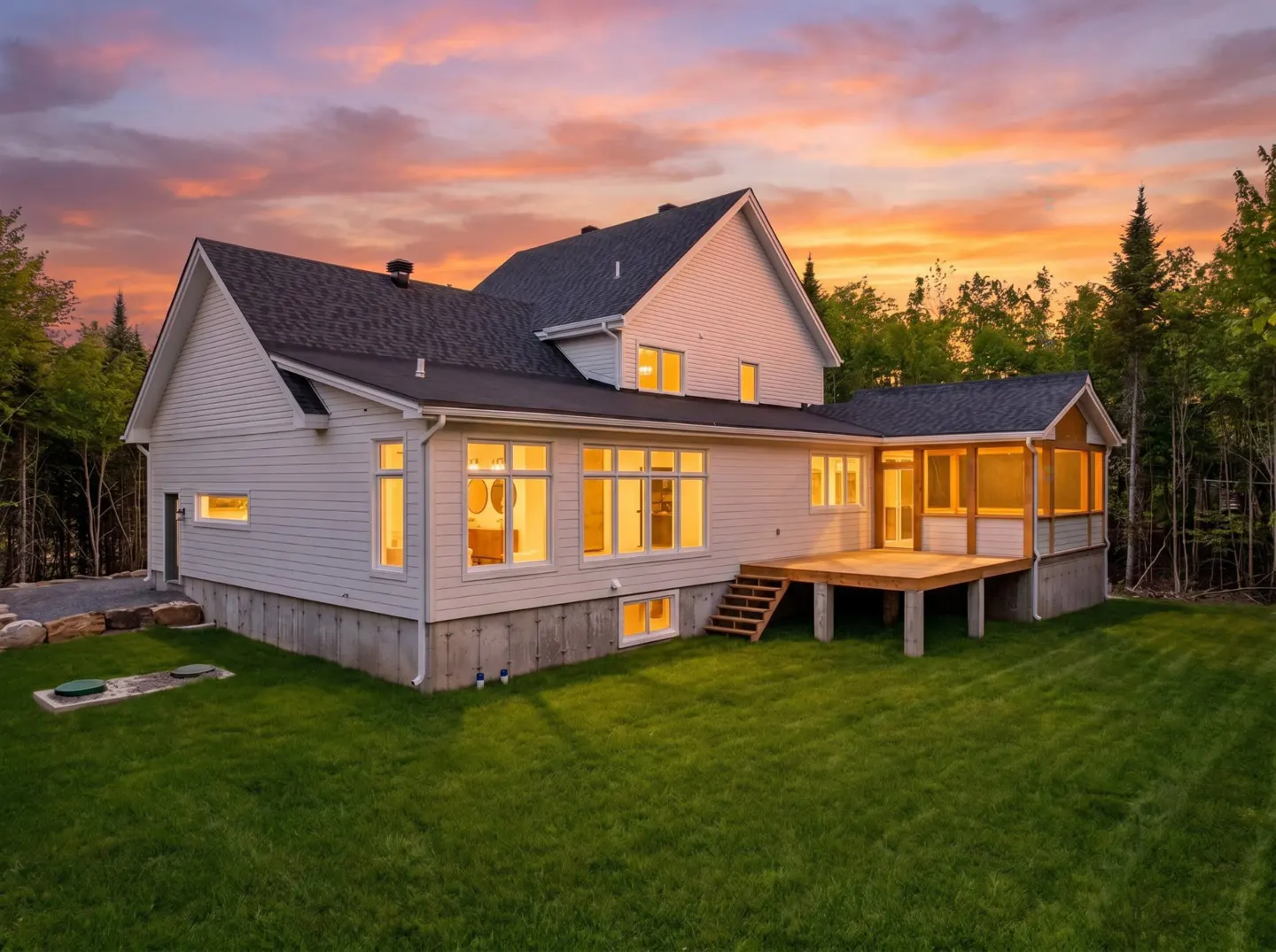 White house with a wooden deck and large windows at sunset, surrounded by a green lawn and forest.
