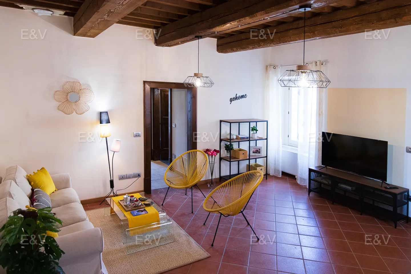 Living room with exposed wood beams, white walls, and terracotta tile floor. Yellow chairs and accents add pops of color. A TV and shelving unit are visible.