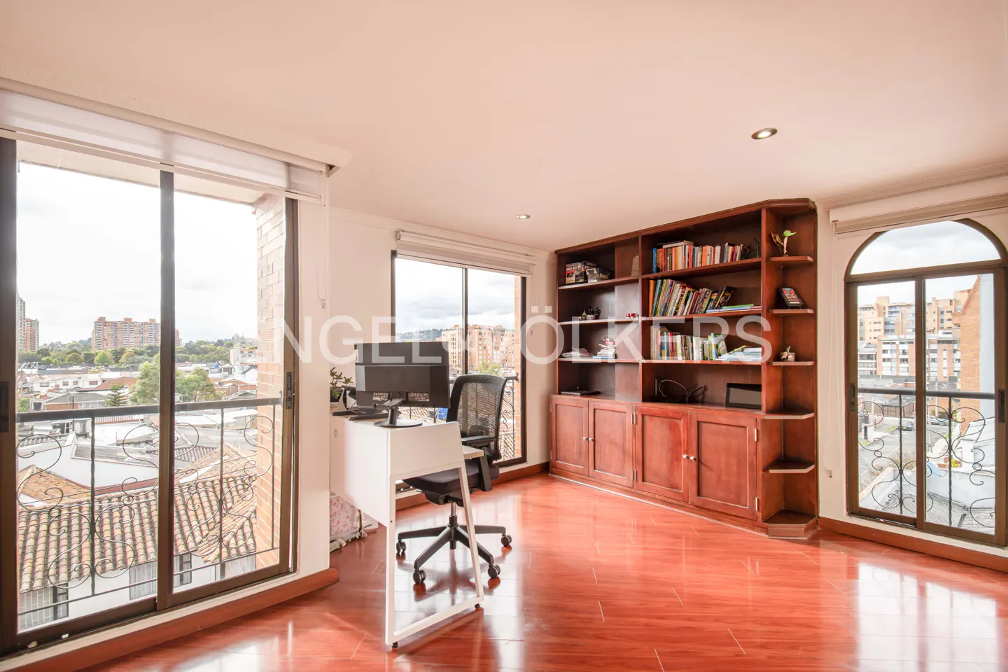 A bright room with a desk, computer, chair, and a large wooden bookcase filled with books. Balcony with city view.