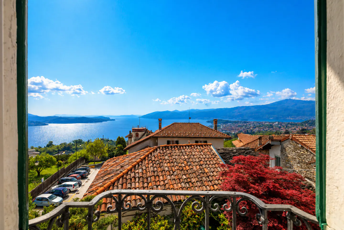 View from a balcony overlooking terracotta rooftops, a blue lake, and mountains under a bright blue sky.