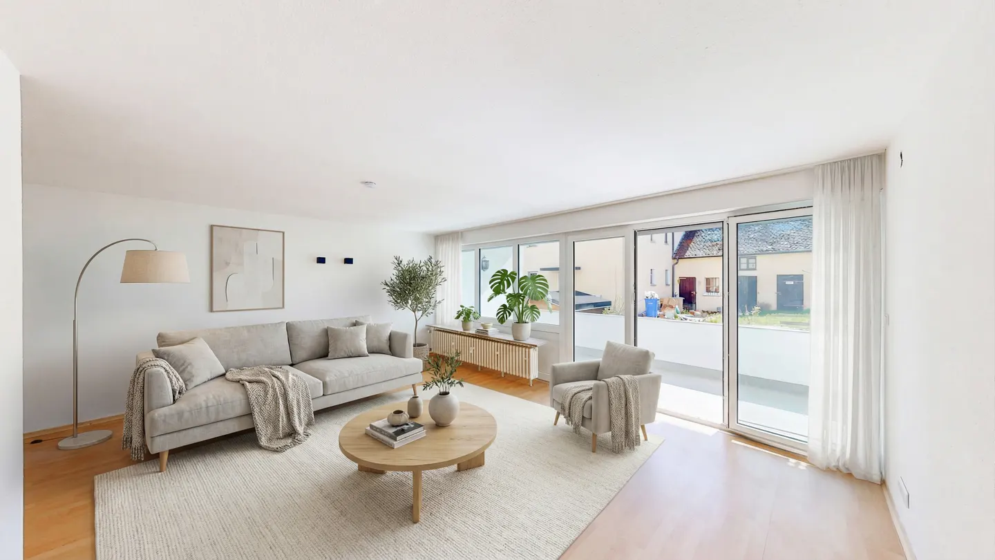 Bright living room with beige sofa, chair, and round wood table on a cream rug. Large windows overlook a patio.