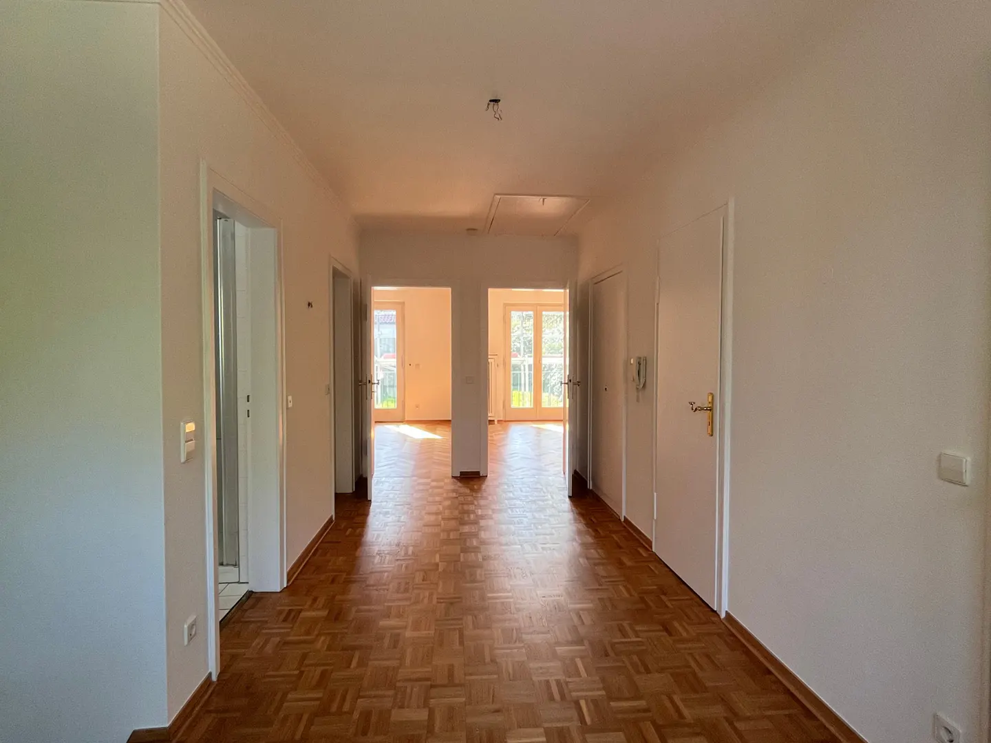 Hallway with parquet flooring, white walls, and doors. Light streams in from the end, illuminating the space.