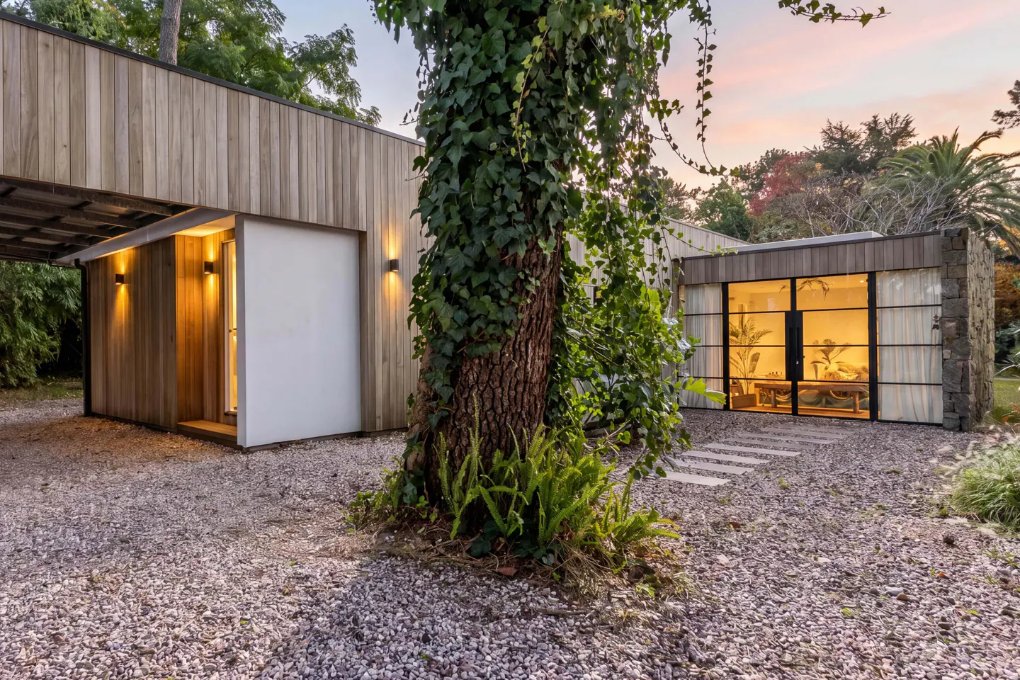 Modern home exterior with wood siding, stone accents, and large windows. A tree covered in ivy stands in the gravel yard.