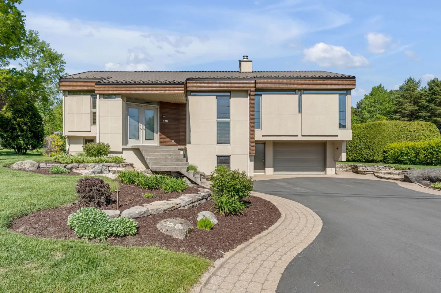 Modern two-story home with beige siding, wood accents, a front staircase, and an attached garage on a landscaped lot.