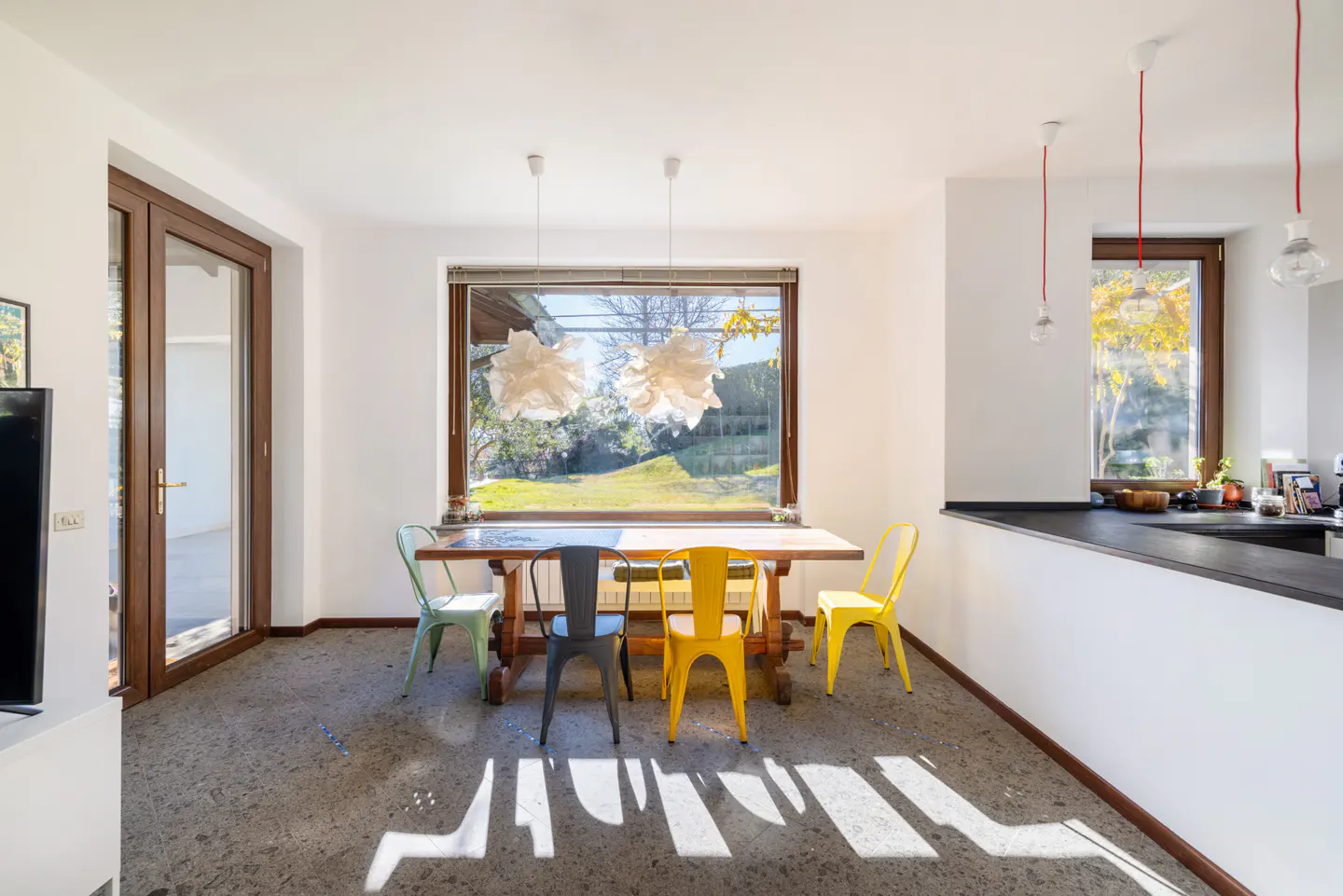 Bright dining area with a wooden table, colorful chairs, and large windows overlooking a green lawn. Two pendant lights hang above the table.