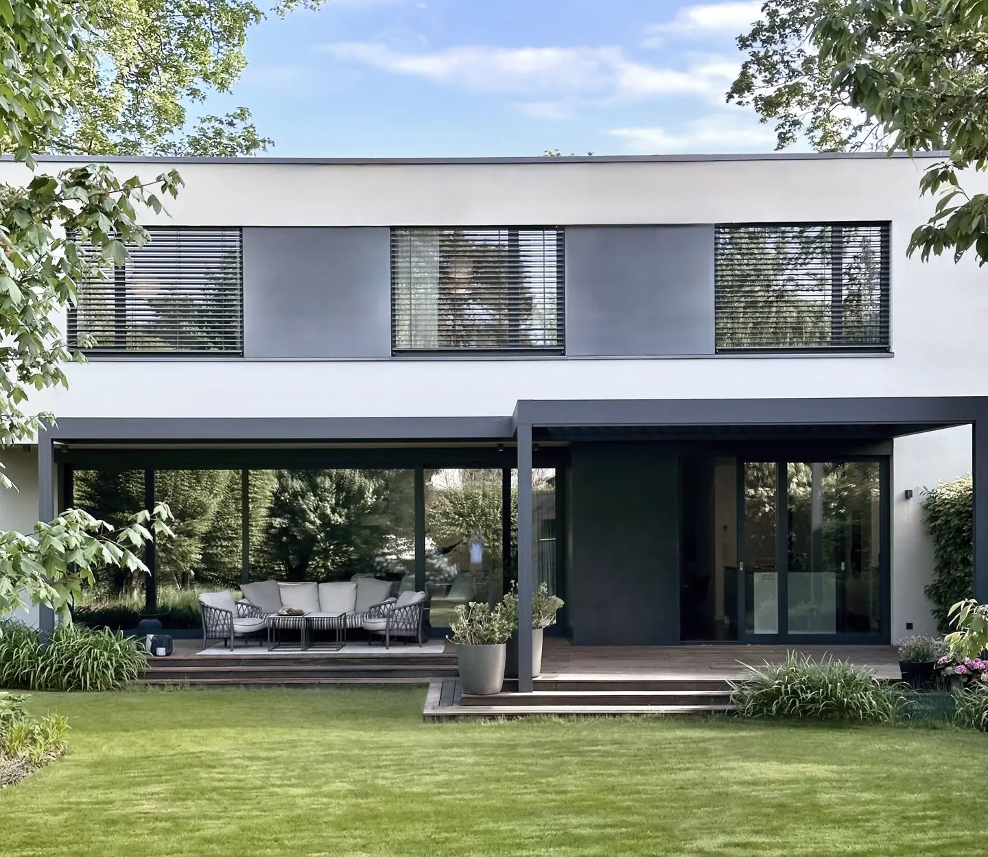 Modern two-story house with a white facade, gray trim, and large windows overlooking a green lawn and patio with outdoor furniture.