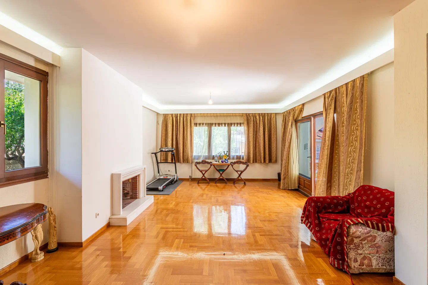 Spacious living room with hardwood floors, a fireplace, a treadmill, a red armchair, and gold curtains over large windows.