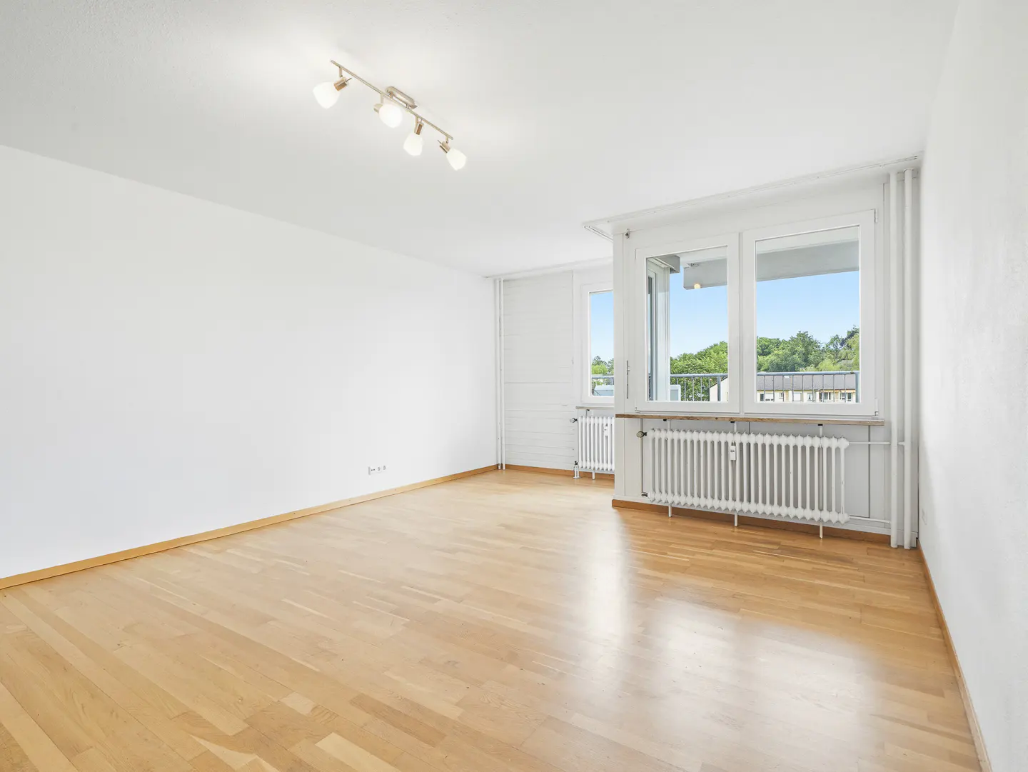 Bright, empty room with light wood floors, white walls, and a multi-bulb ceiling light. Windows show trees outside.