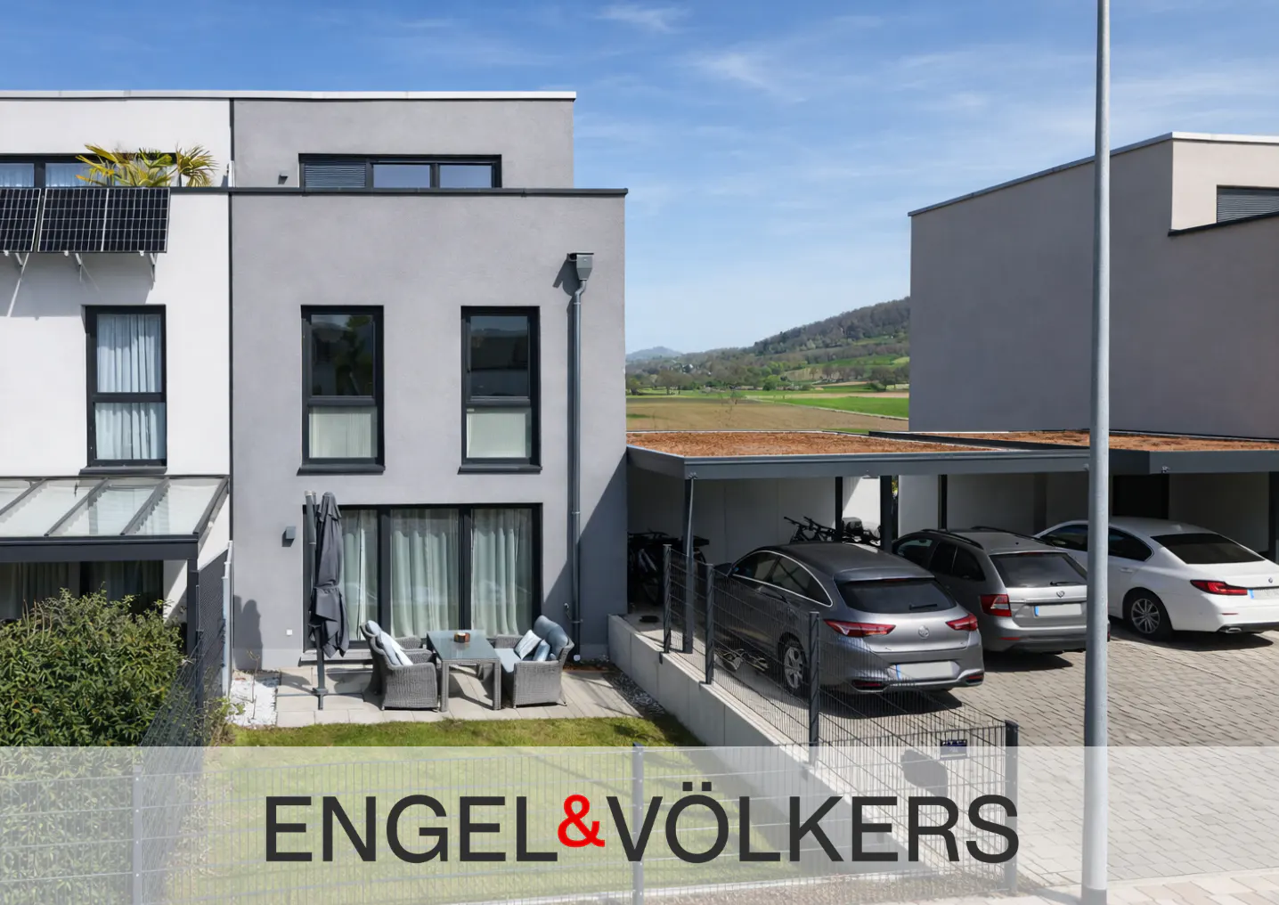 Modern gray townhouses with a patio, outdoor seating, and a carport containing three parked cars under a sunny blue sky.
