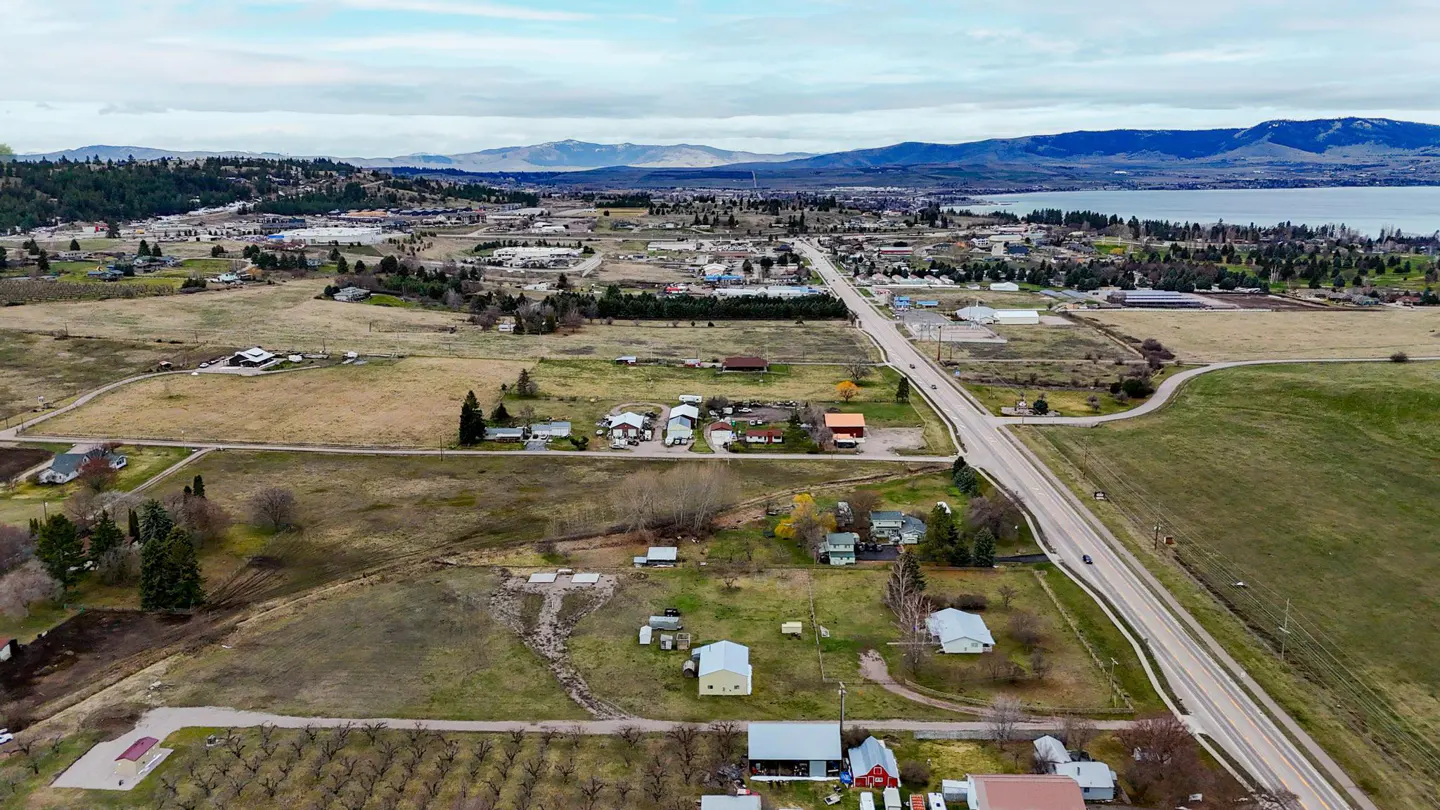 Aerial view of a rural landscape with houses, fields, and a road leading to a town near a lake and mountains.