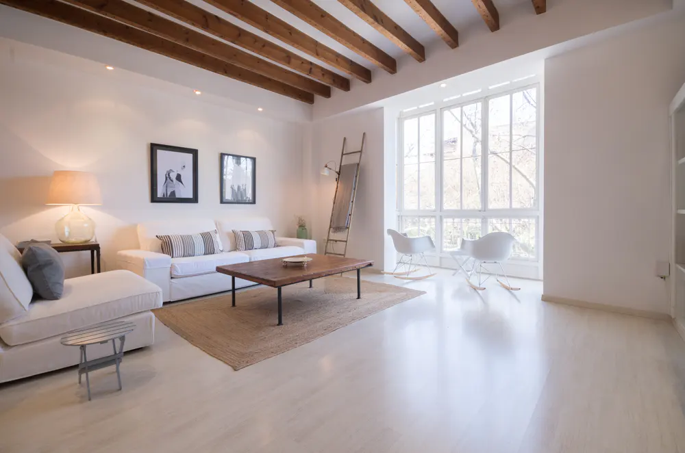 Bright living room with white walls, wooden ceiling beams, and large windows. White sofas, a wooden coffee table, and two white rocking chairs furnish the space.