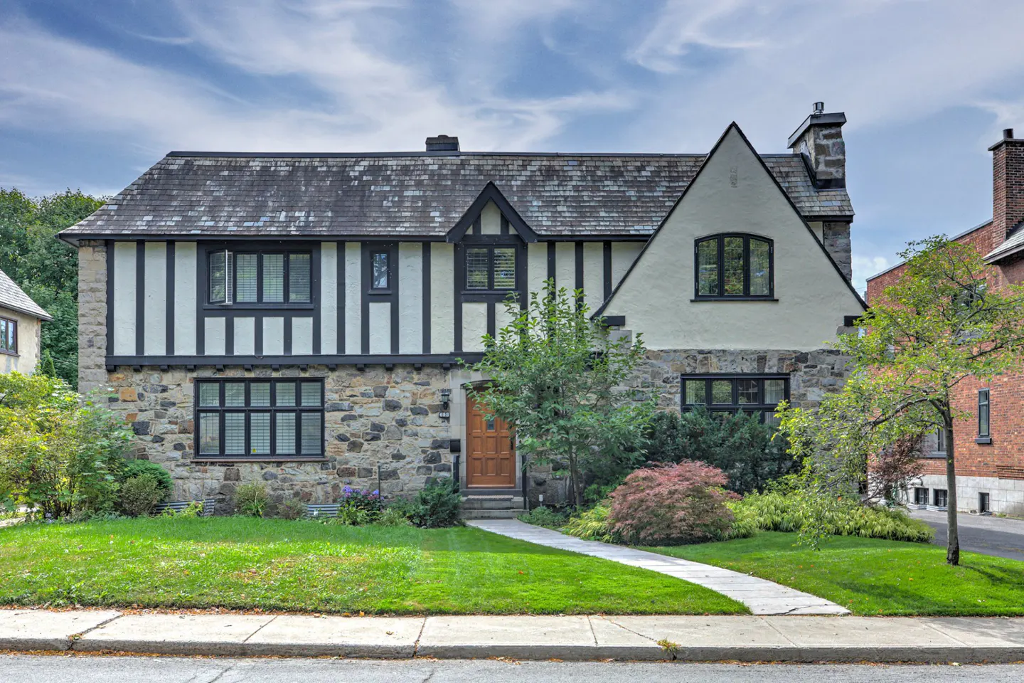 Two-story Tudor-style house with stone and stucco exterior, black trim, and a gray shingle roof. A walkway leads to the wooden front door.