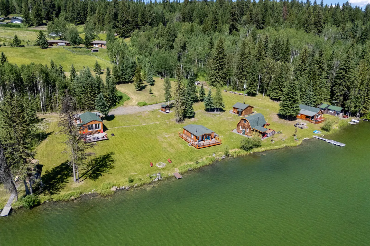 Aerial view of lakeside cabins with green roofs, surrounded by lush trees and green grass. A dock extends into the green water.