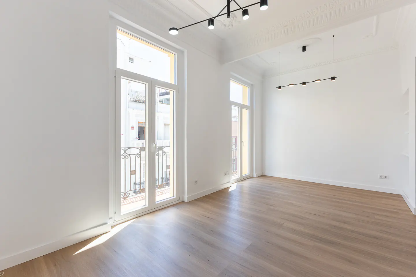 Bright, empty room with light wood floors, white walls, and two white-framed glass doors with iron railings leading to balconies. Modern black light fixtures hang from the ornate white ceiling.