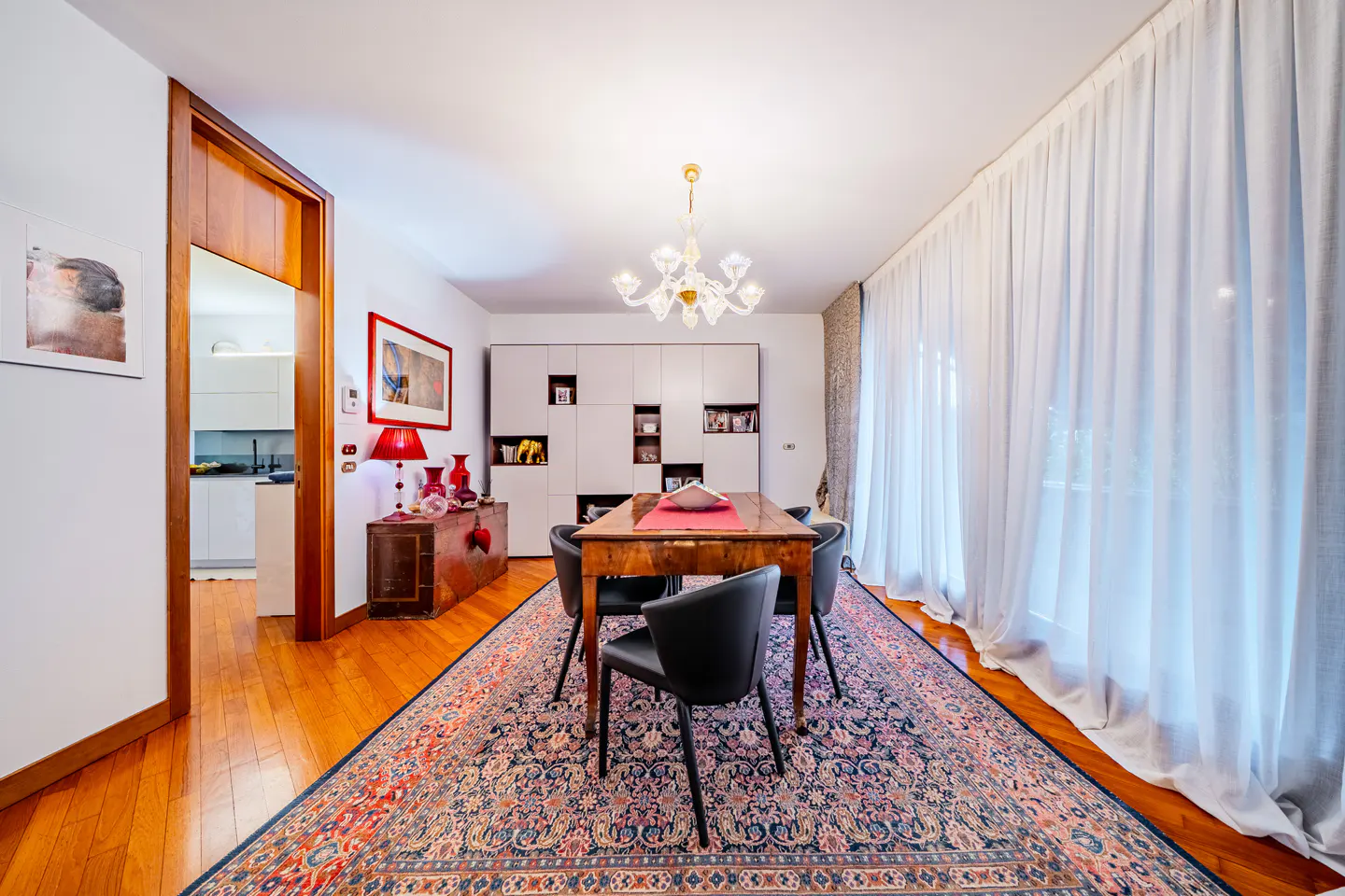A dining room with a wooden table, black chairs, and a colorful rug. White curtains and a chandelier add elegance.