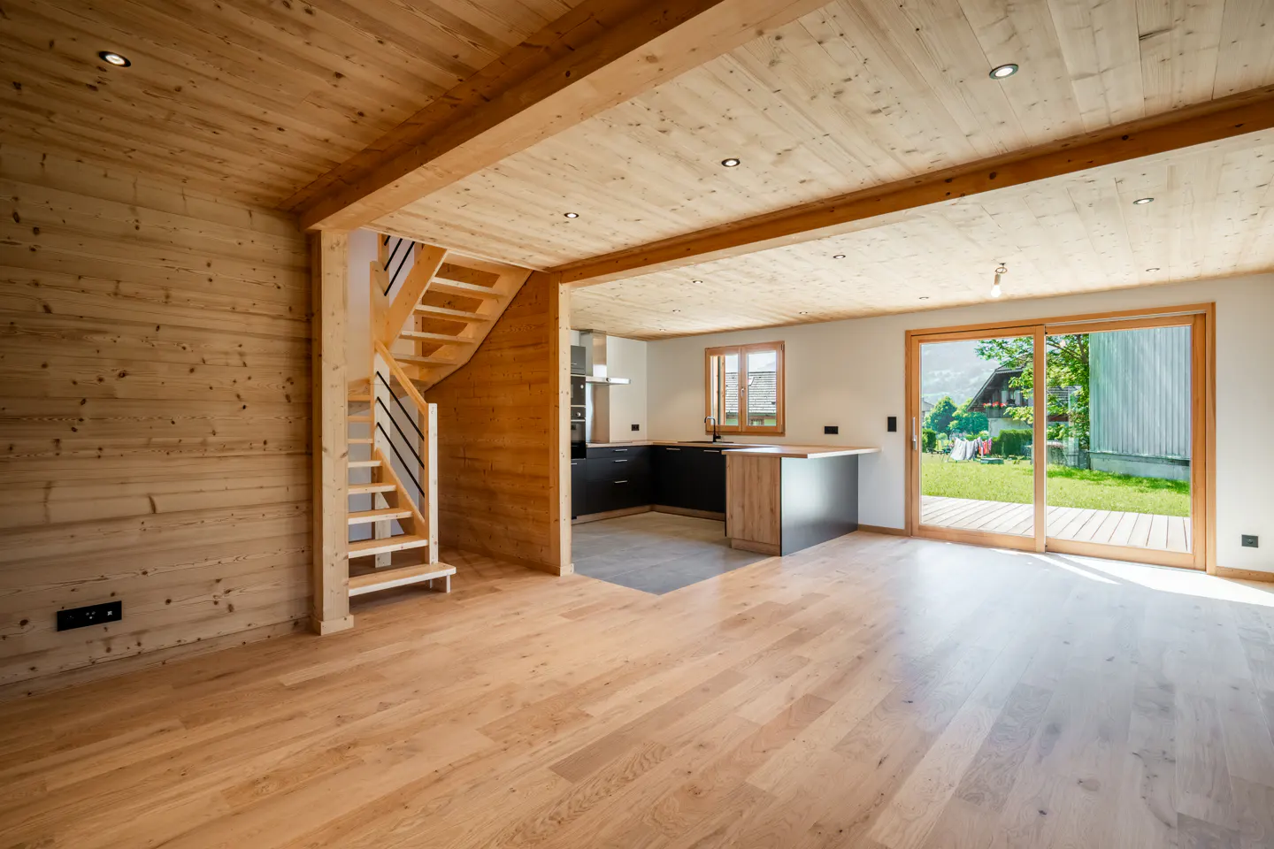 Bright, modern interior with wood walls, floors, and ceiling. A staircase leads up, and a kitchen is visible through an opening. Sliding glass doors open to a green lawn.
