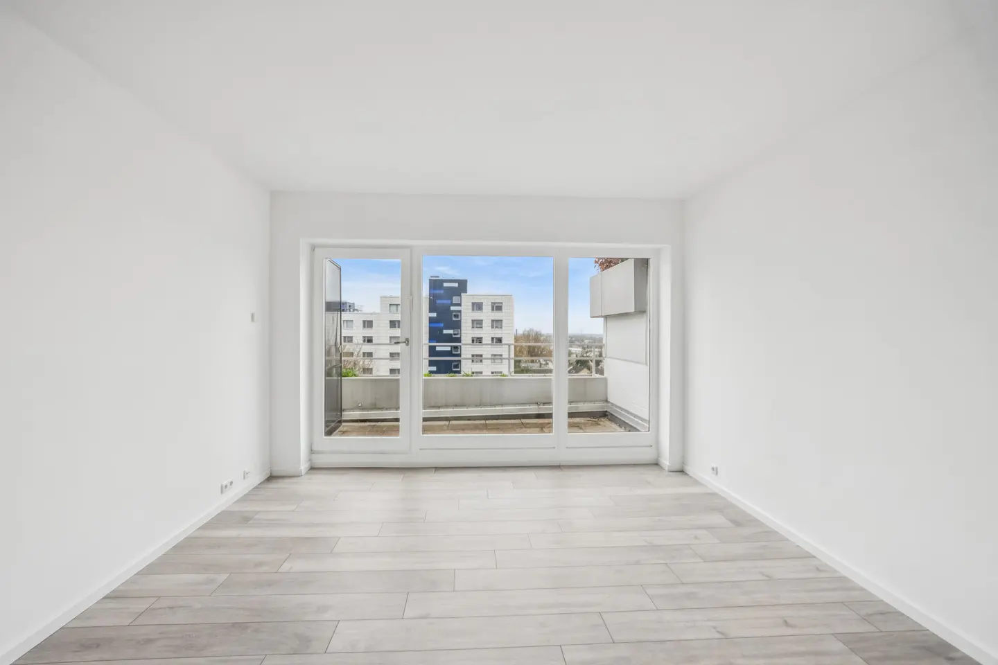 Bright, empty room with white walls and gray wood floors. Large glass doors open to a balcony with a city view.