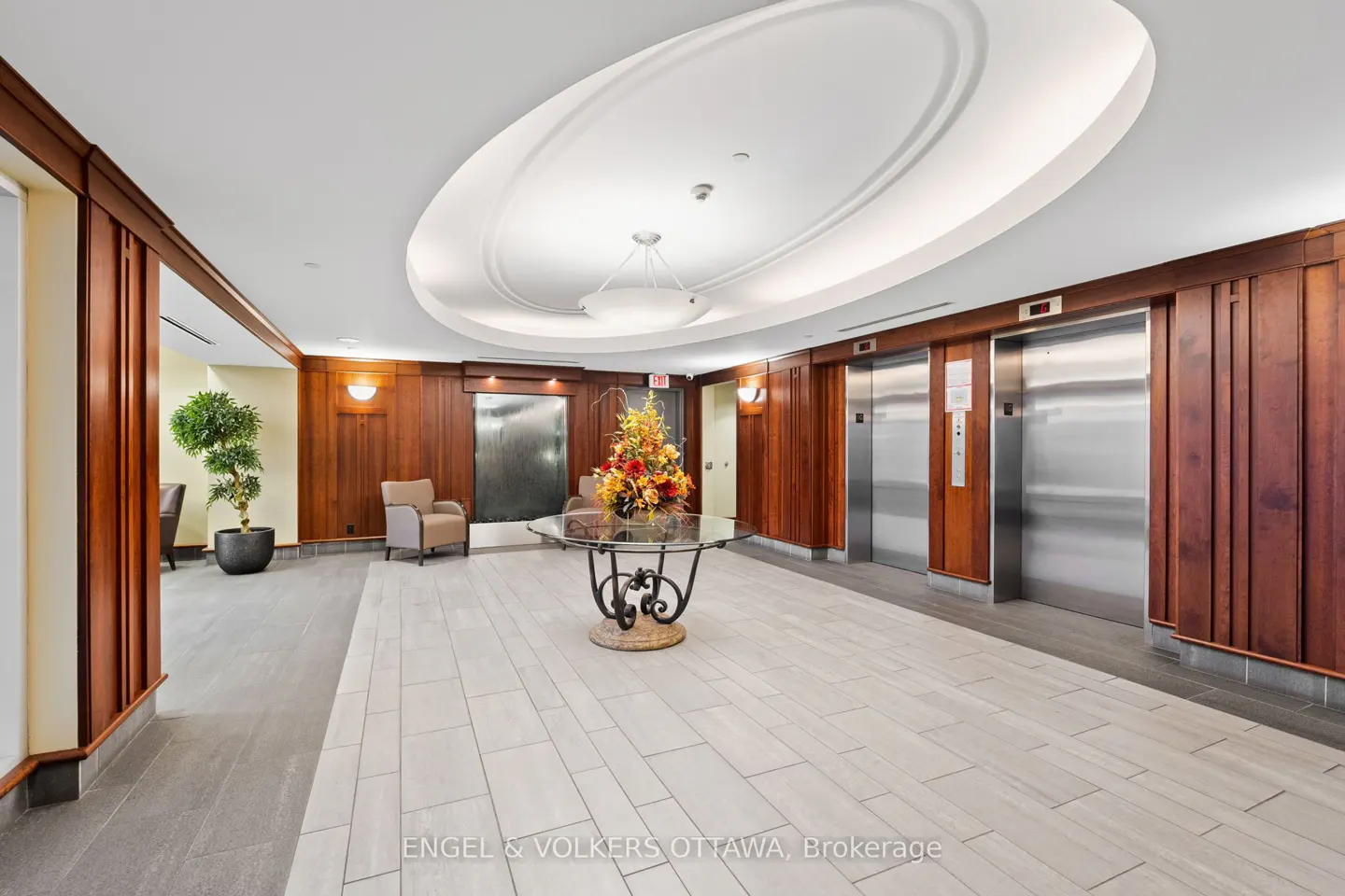 Lobby with tile floor, wood paneling, and two elevators. A flower arrangement sits on a glass table. A water feature is in the background.