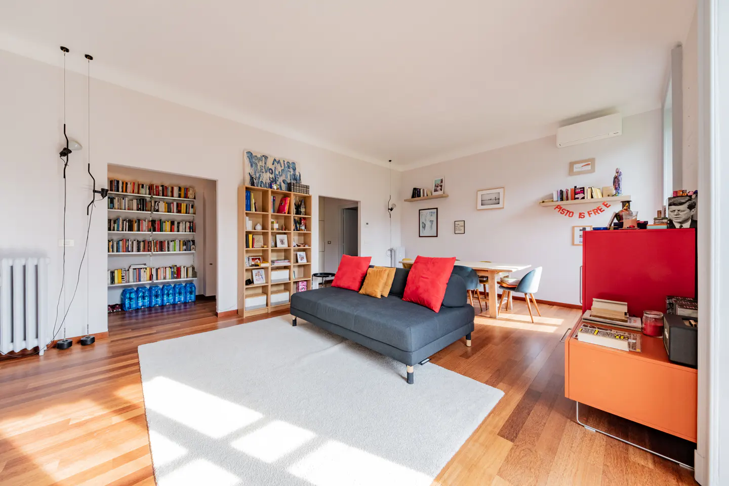 Bright living room with hardwood floors, white rug, and gray sofa with red pillows. Bookshelves and colorful furniture add character.