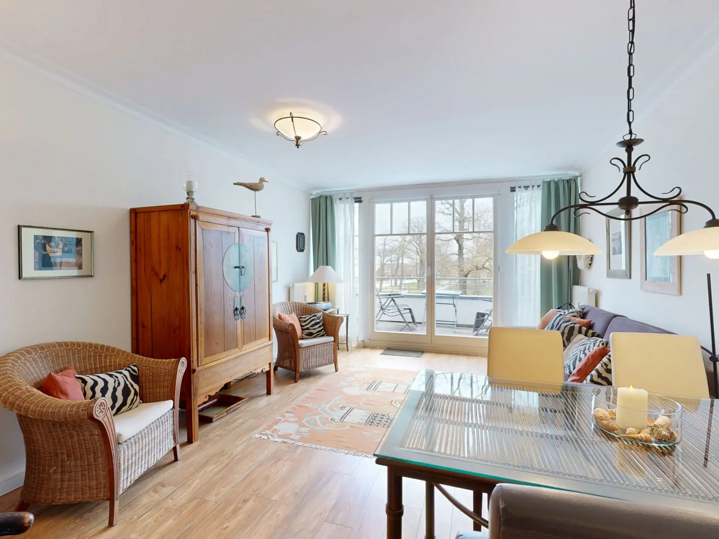 Bright living room with wood floors, wicker chairs, and a glass dining table. A balcony is visible through large sliding glass doors.