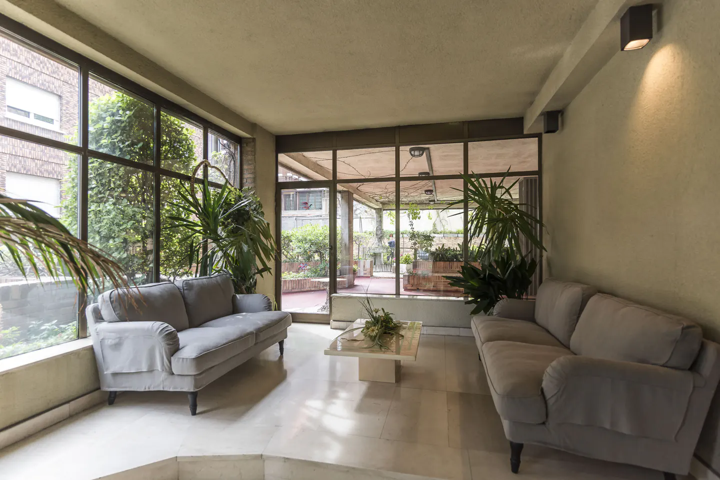 Sunroom with two grey sofas, a central coffee table, and large windows overlooking a garden patio with lush greenery.
