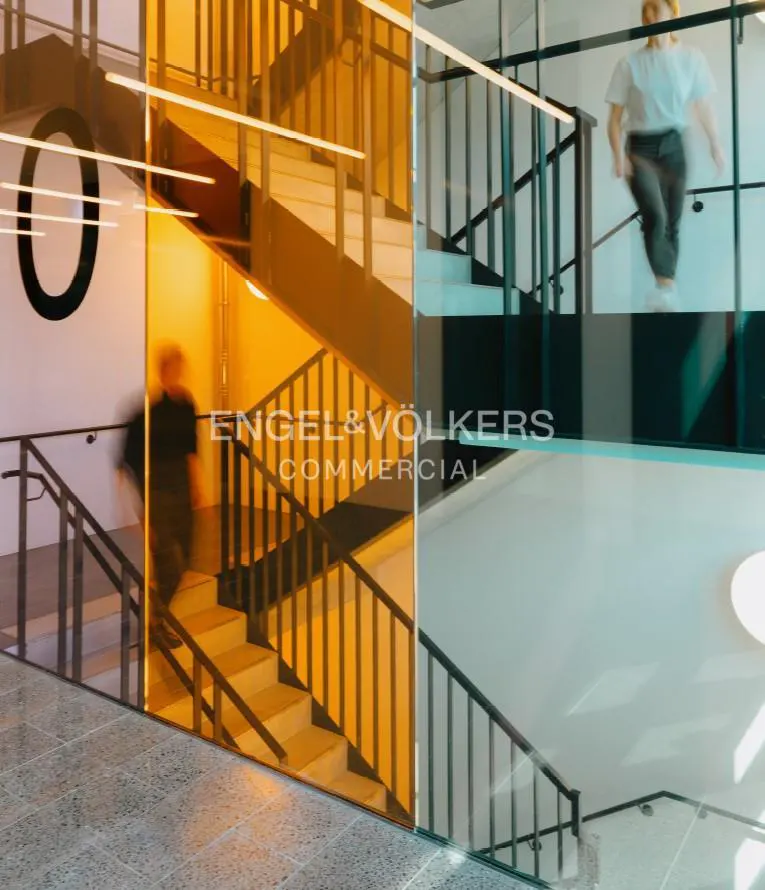 Interior view of Engel & Völkers Commercial office with stairs, people walking, and the company logo on a glass wall.