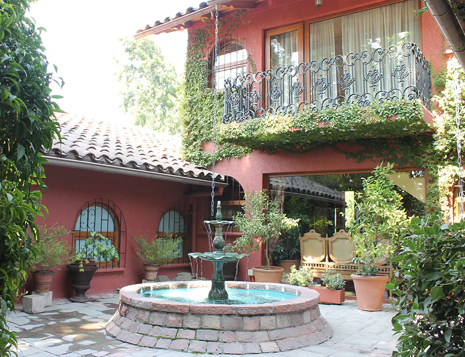 Courtyard view of a two-story red building with a stone fountain, plants, and a wrought iron balcony.