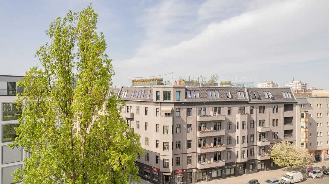 View of a gray apartment building with balconies, street-level shops, and tall green trees against a blue sky.