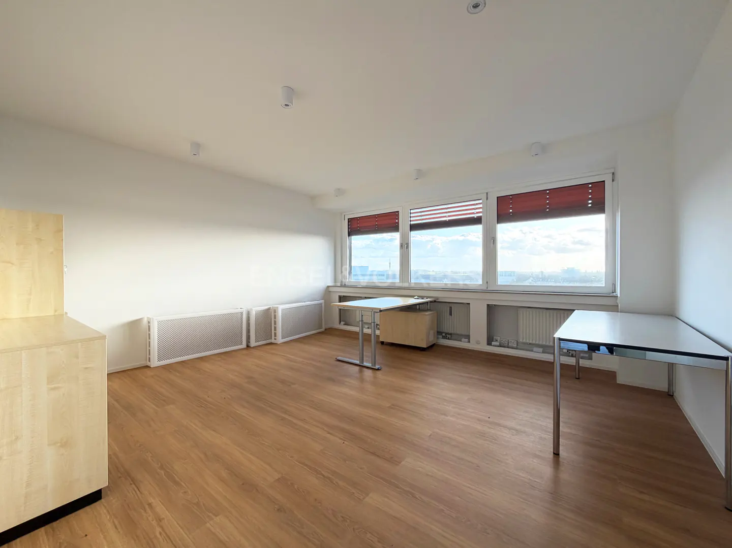 Bright, empty office space with wood floors, white walls, and large windows with red blinds. Two tables and a light wood cabinet are visible.
