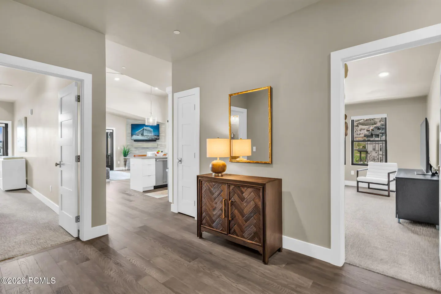 Hallway with wood floors, beige walls, and white trim. A wooden cabinet with lamps and a gold-framed mirror sits against the wall. Open doorways lead to other rooms.