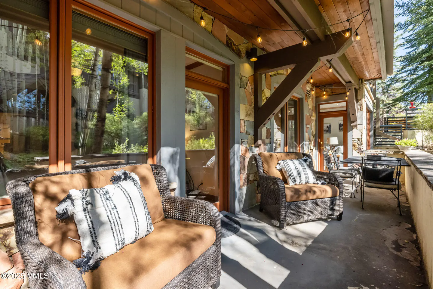 Outdoor patio with wicker chairs, brown cushions, and striped pillows. String lights hang above a stone wall and wood ceiling.