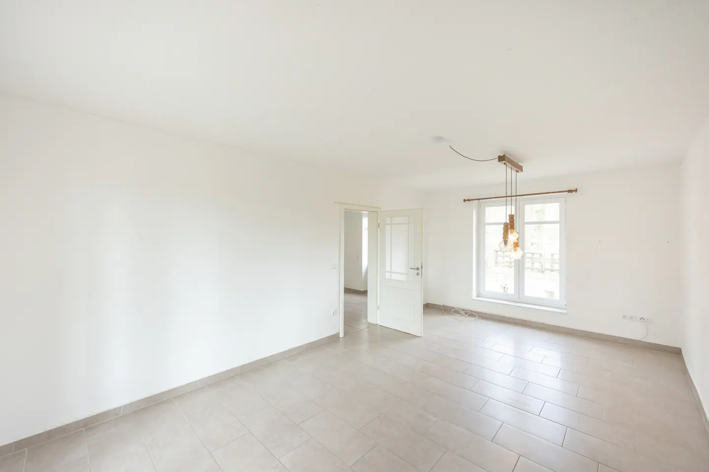 Bright, empty room with white walls, tile floor, and a window with hanging lights. A white door is slightly ajar.