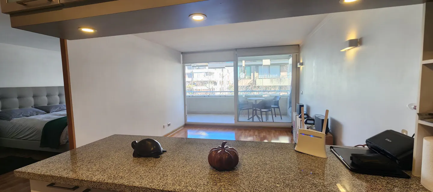 A modern apartment interior with a granite countertop, turtle and pumpkin decor, and a balcony with outdoor seating. A bedroom is visible in the background.