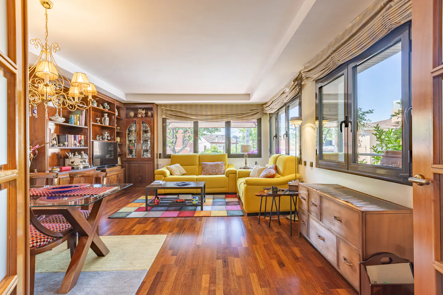 Bright living room with hardwood floors, yellow sofa, colorful rug, and built-in wooden shelves. Large windows offer natural light.