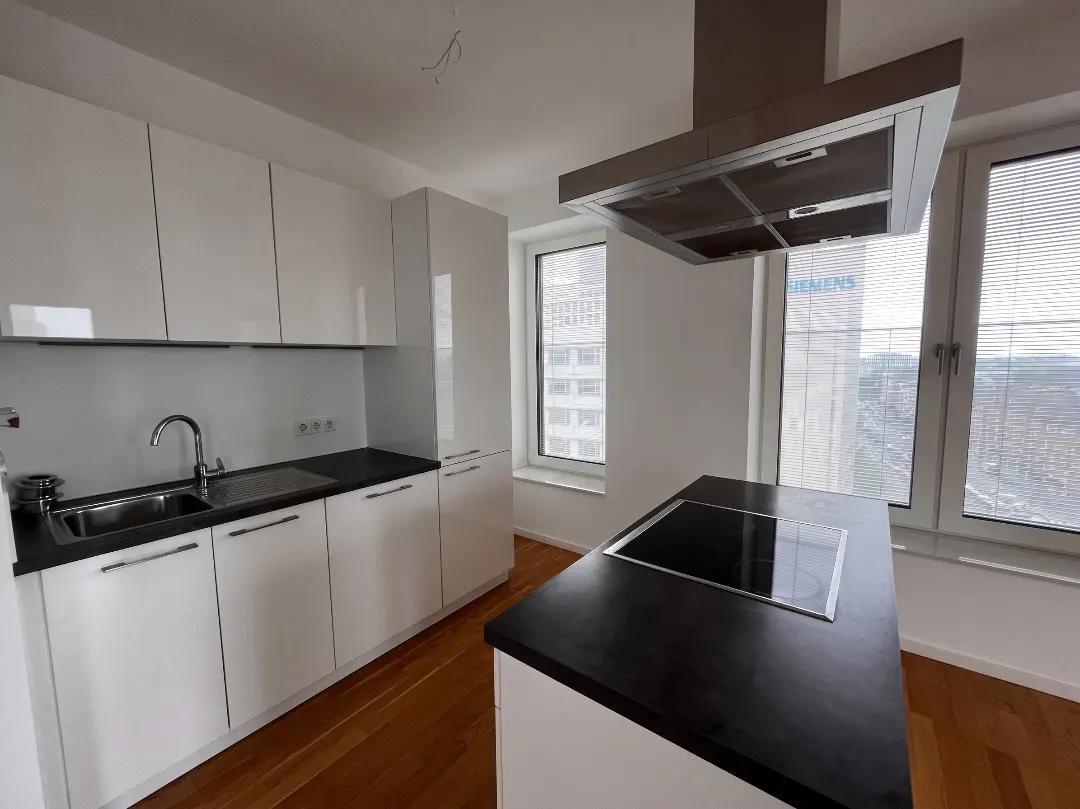 A modern kitchen with white cabinets, black countertops, and a stainless steel range hood. A kitchen island with a cooktop stands in the center. Windows offer a city view.