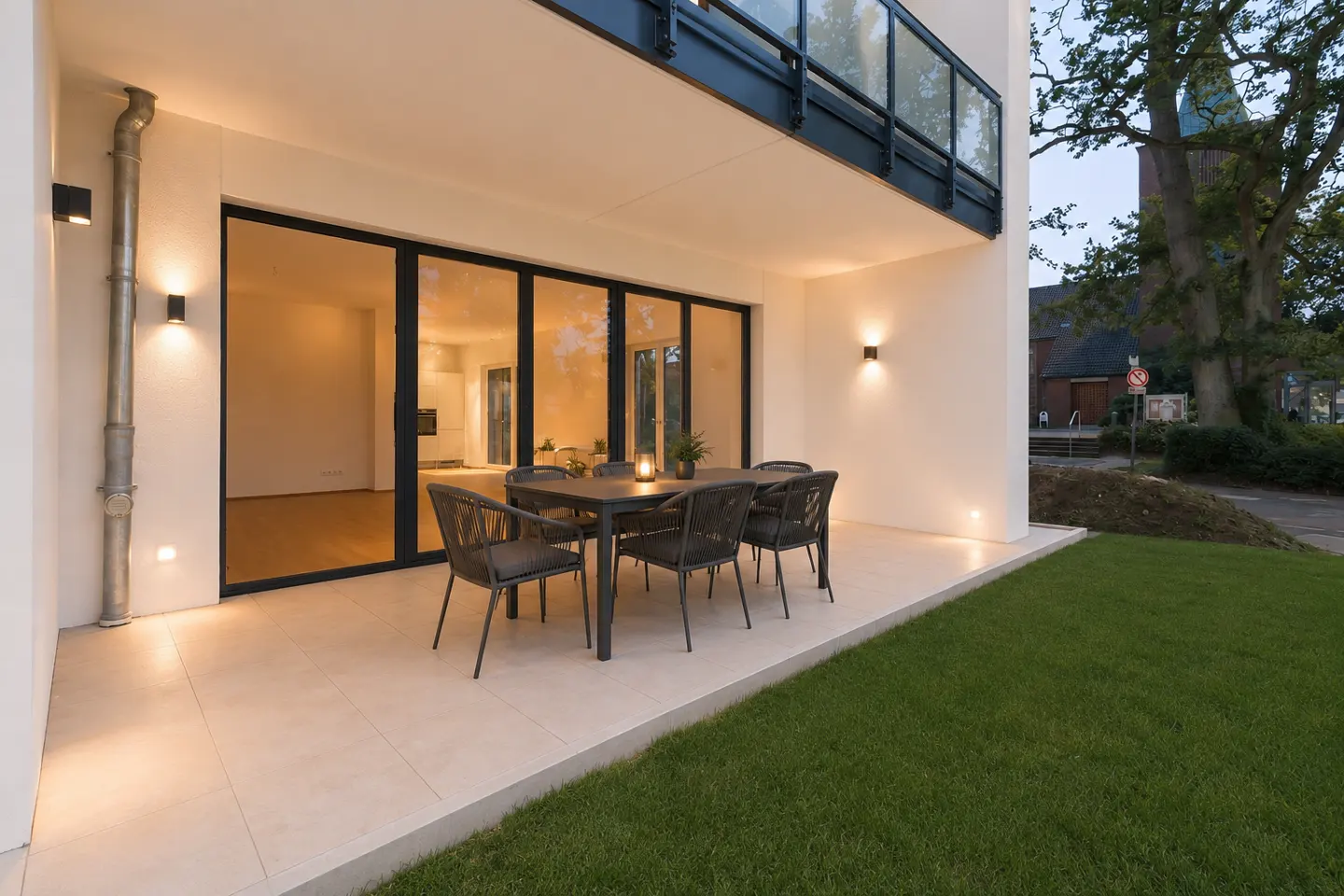 Modern home exterior with patio table and chairs. Large glass doors lead inside. Green lawn and trees in the background.