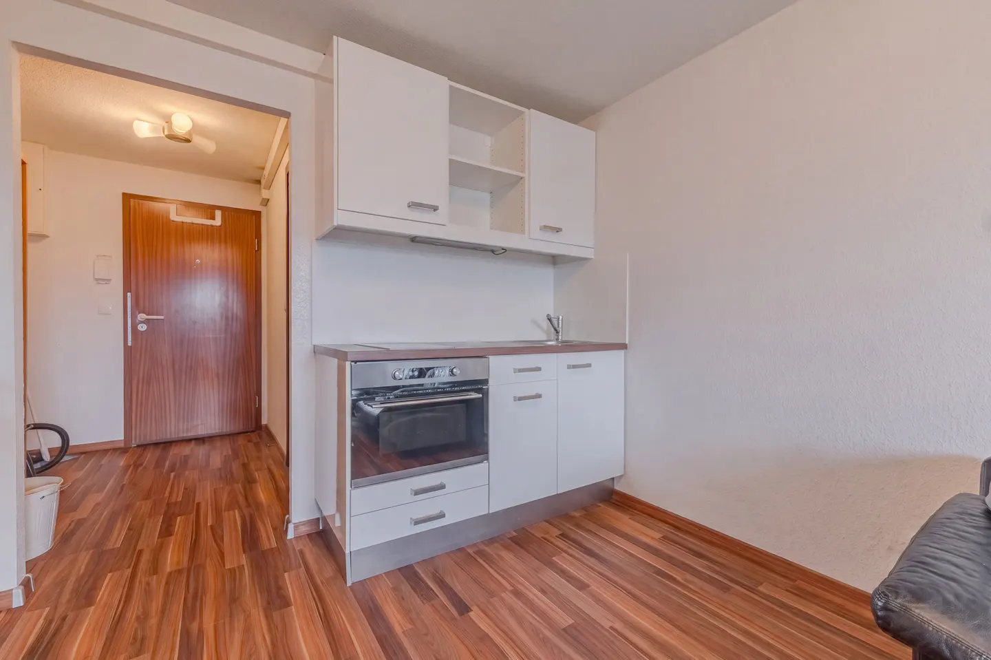 A small kitchen with white cabinets, a stainless steel oven, and wood floors. A brown door is visible in the background.