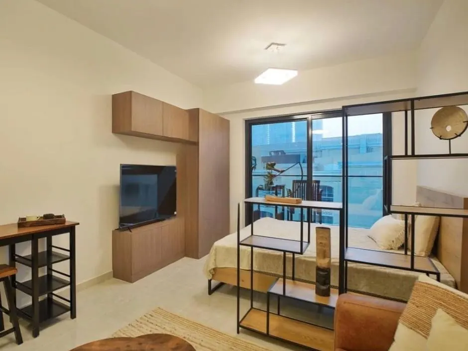 A modern studio apartment with a bed, TV, and shelving. The room has a neutral color palette with wood accents and a large window.
