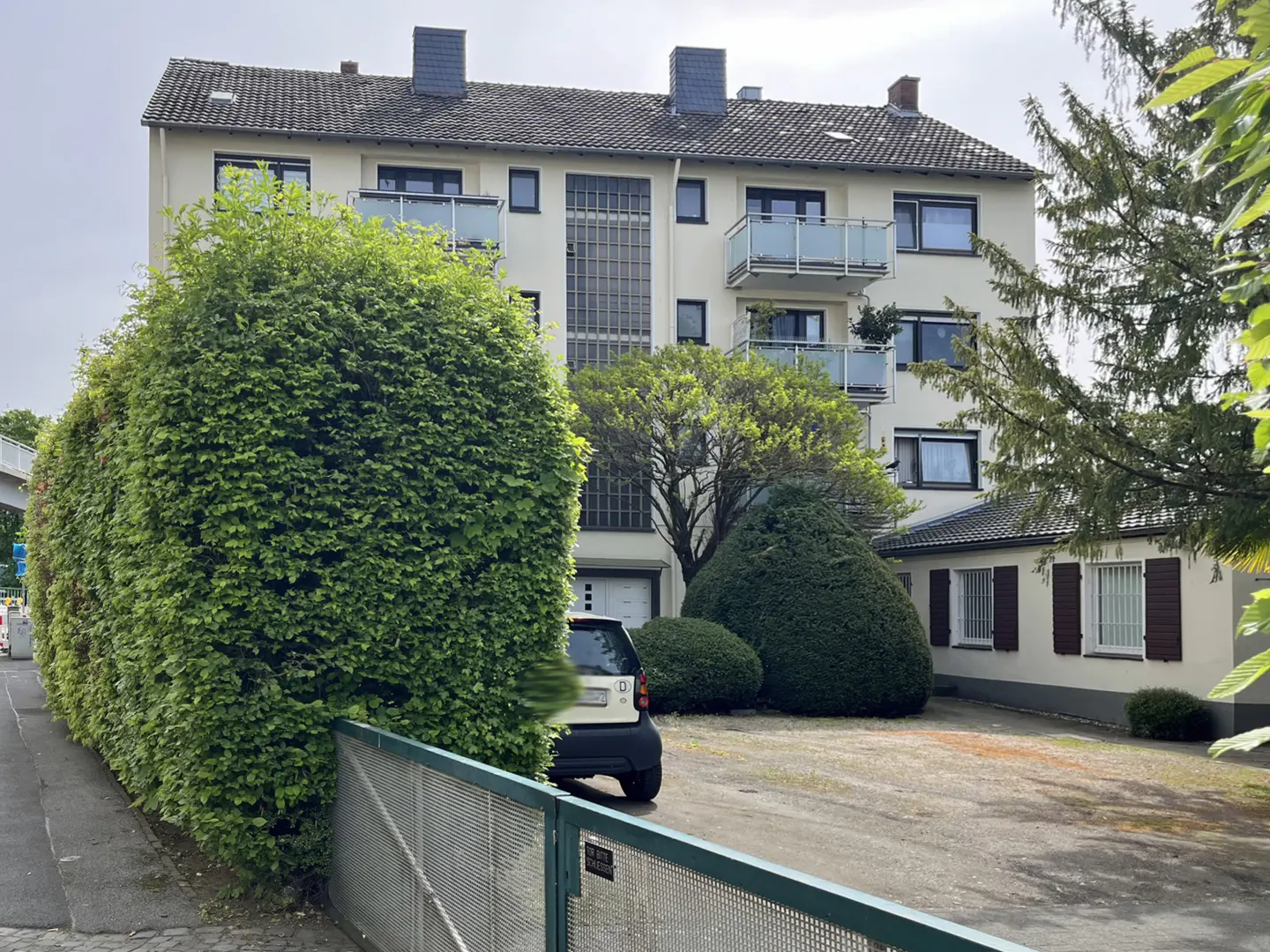 A multi-story cream apartment building with balconies, a gravel parking area, and large, manicured green hedges.