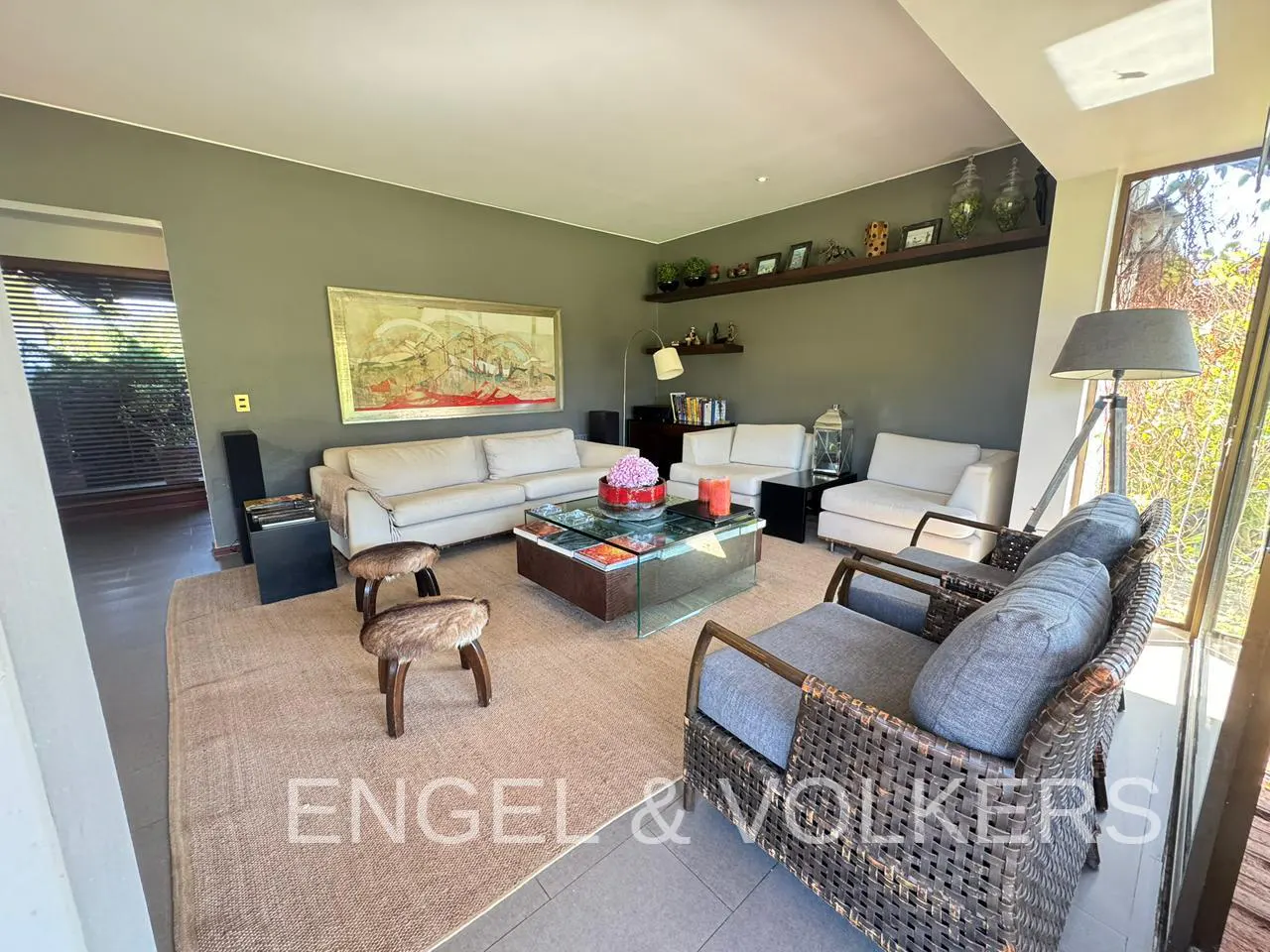 A living room with white sofas, a glass coffee table, and wicker chairs on a tan rug. An abstract painting hangs on the gray wall.